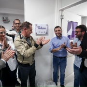 A imagem mostra um grupo de pessoas em um ambiente interno, comemorando a inauguração de um espaço chamado "Ser Mulher RS", conforme indicado pela placa na parede. A placa tem o logotipo do governo do Rio Grande do Sul e de outros órgãos públicos.
No centro, um homem de camisa azul clara e calça jeans está sorrindo e aplaudindo, cercado por outras pessoas que também aplaudem e sorriem. À esquerda da imagem, há uma mulher com óculos de armação azul e cabelo preso, usando um blazer branco. Outras pessoas ao redor vestem roupas sociais, como ternos, camisas e blazers. A parede ao fundo é branca, e há uma porta aberta com um cartaz roxo que parece ilustrar o perfil de uma mulher.
O clima da cena é de celebração e realização, sugerindo a inauguração de um serviço ou espaço voltado ao público feminino no estado do Rio Grande do Sul.