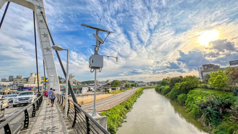 Fotografia de uma ponte urbana com passarela para pedestres, onde algumas pessoas caminham. À esquerda, há carros circulando na pista de veículos. Na lateral da passarela, está instalada uma estrutura metálica com placas solares e equipamentos de monitoramento. Ao fundo, vê-se um rio margeado por vegetação verde de um lado e por uma via asfaltada do outro. No horizonte aparecem prédios e construções comerciais da cidade. O céu está parcialmente nublado, com o sol brilhando próximo ao entardecer, criando tons dourados e azuis na cena.