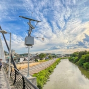 Fotografia de uma ponte urbana com passarela para pedestres, onde algumas pessoas caminham. À esquerda, há carros circulando na pista de veículos. Na lateral da passarela, está instalada uma estrutura metálica com placas solares e equipamentos de monitoramento. Ao fundo, vê-se um rio margeado por vegetação verde de um lado e por uma via asfaltada do outro. No horizonte aparecem prédios e construções comerciais da cidade. O céu está parcialmente nublado, com o sol brilhando próximo ao entardecer, criando tons dourados e azuis na cena.