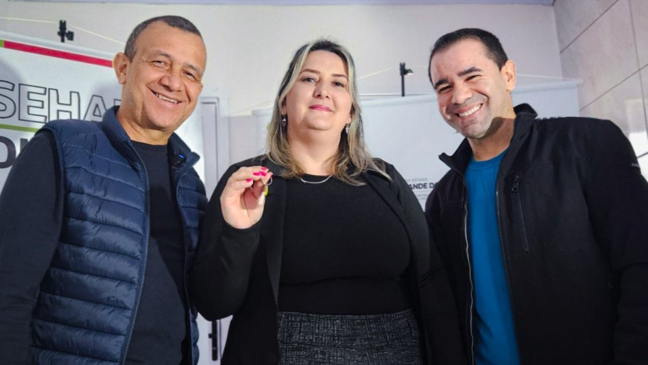 A imagem mostra três pessoas sorrindo e posando para a foto em um ambiente interno. Ao centro está uma mulher de cabelos loiros e lisos, vestindo uma blusa preta e saia cinza, segurando uma chave com a mão direita. À esquerda, um homem de pele morena, com cabelo curto, usando uma jaqueta acolchoada azul-marinho e camiseta preta. À direita, outro homem também de pele morena, com cabelo curto, vestindo uma jaqueta preta sobre uma camiseta azul. Ao fundo, há uma faixa com parte de um texto visível que parece estar relacionada a um evento ou entrega, possivelmente institucional ou governamental, sugerido pelo estilo do ambiente e a presença da chave, que pode simbolizar a entrega de um imóvel ou veículo.