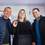 A imagem mostra três pessoas sorrindo e posando para a foto em um ambiente interno. Ao centro está uma mulher de cabelos loiros e lisos, vestindo uma blusa preta e saia cinza, segurando uma chave com a mão direita. À esquerda, um homem de pele morena, com cabelo curto, usando uma jaqueta acolchoada azul-marinho e camiseta preta. À direita, outro homem também de pele morena, com cabelo curto, vestindo uma jaqueta preta sobre uma camiseta azul. Ao fundo, há uma faixa com parte de um texto visível que parece estar relacionada a um evento ou entrega, possivelmente institucional ou governamental, sugerido pelo estilo do ambiente e a presença da chave, que pode simbolizar a entrega de um imóvel ou veículo.