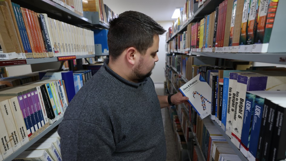 Nesta imagem, um homem está em uma biblioteca, em frente a prateleiras cheias de livros técnicos e acadêmicos. Ele segura um exemplar intitulado “Java Prático”, indicando o foco em tecnologia e programação. 