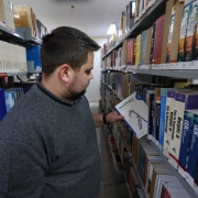 Nesta imagem, um homem está em uma biblioteca, em frente a prateleiras cheias de livros técnicos e acadêmicos. Ele segura um exemplar intitulado “Java Prático”, indicando o foco em tecnologia e programação. 