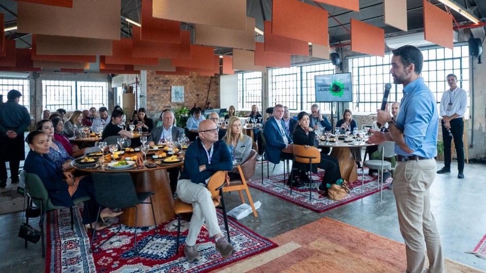 A imagem retrata um encontro formal em um salão com grandes janelas e decoração contemporânea. Pessoas estão sentadas ao redor de mesas redondas com pratos e taças, indicando um almoço ou jantar corporativo. No primeiro plano, o governador está em pé com microfone, fazendo uma apresentação.