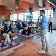 A imagem retrata um encontro formal em um salão com grandes janelas e decoração contemporânea. Pessoas estão sentadas ao redor de mesas redondas com pratos e taças, indicando um almoço ou jantar corporativo. No primeiro plano, o governador está em pé com microfone, fazendo uma apresentação.
