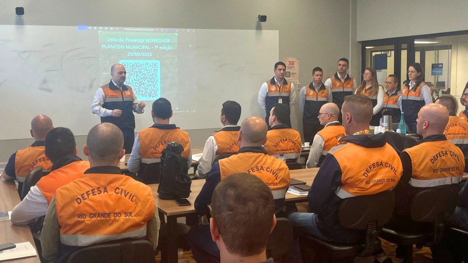 Foto mostra Coronel Boeira em pé falando diante de sala cheia de participantes usando coletes laranja da Defesa Civil do Rio Grande do Sul. Atrás dele, outros integrantes da equipe estão alinhados de frente para o público. No fundo, um projetor exibe a lista de presença do workshop, com QR Code visível.