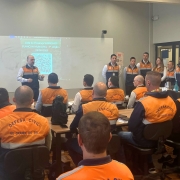 Foto mostra Coronel Boeira em pé falando diante de sala cheia de participantes usando coletes laranja da Defesa Civil do Rio Grande do Sul. Atrás dele, outros integrantes da equipe estão alinhados de frente para o público. No fundo, um projetor exibe a lista de presença do workshop, com QR Code visível.