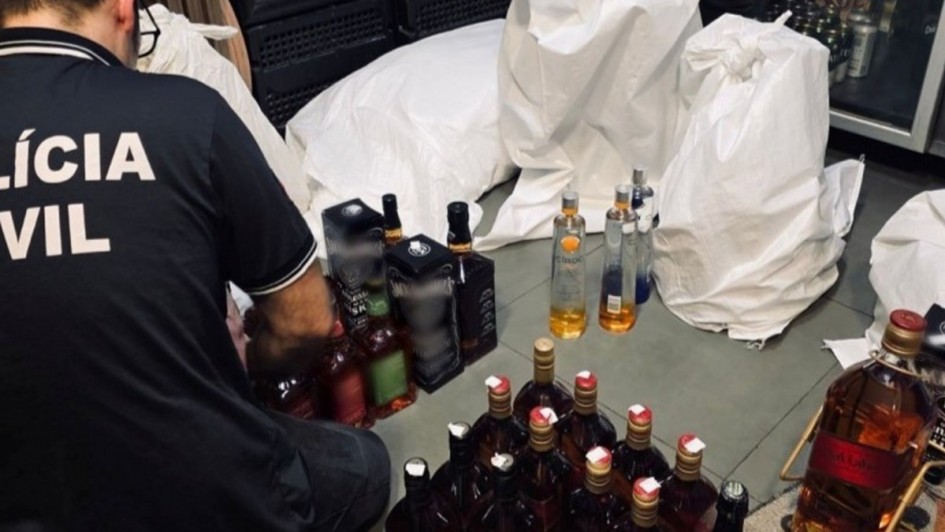 Na foto, um agente da Polícia Civil está agachado e organiza diversas garrafas de bebidas alcoólicas, posicionadas em fileiras sobre o chão. Ao lado, há sacos brancos grandes e refrigeradores cheios de produtos.