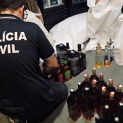 Na foto, um agente da Polícia Civil está agachado e organiza diversas garrafas de bebidas alcoólicas, posicionadas em fileiras sobre o chão. Ao lado, há sacos brancos grandes e refrigeradores cheios de produtos.