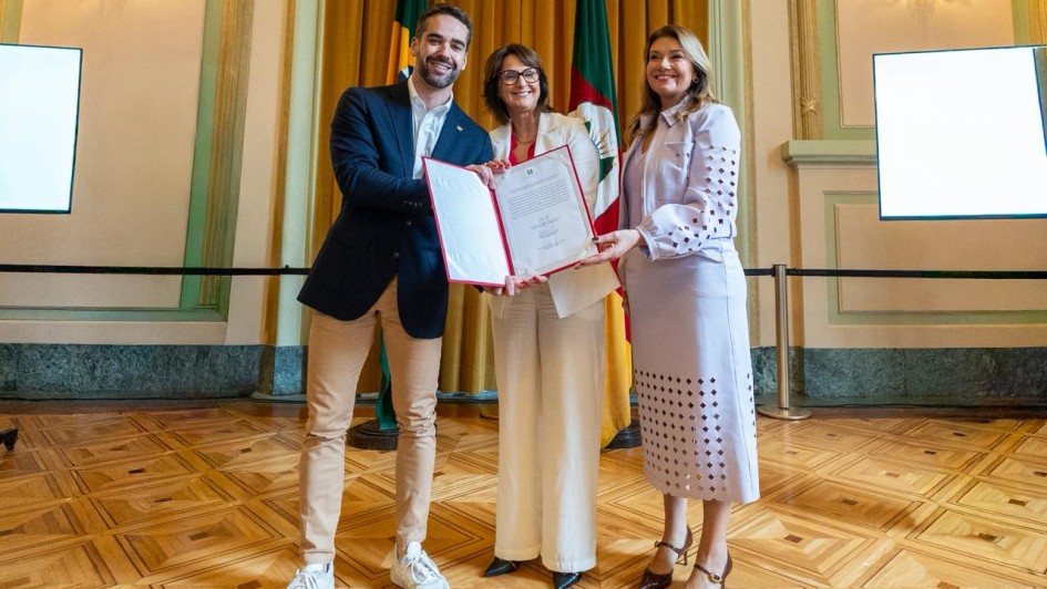 Em um salão com cortinas douradas e piso de madeira clara, três pessoas posam sorrindo para a foto enquanto seguram, juntas, um documento aberto com moldura vermelha. À esquerda, um homem de barba curta, vestindo blazer azul-marinho, calça bege e tênis brancos. Ao centro, uma mulher de cabelos curtos e óculos, trajando conjunto branco, segura o documento. À direita, outra mulher, de cabelos castanhos claros, usa um conjunto lilás com detalhes vazados e sapatos marrons de salto. Ao fundo, aparecem as bandeiras do Brasil e do Rio Grande do Sul.