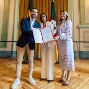 Em um salão com cortinas douradas e piso de madeira clara, três pessoas posam sorrindo para a foto enquanto seguram, juntas, um documento aberto com moldura vermelha. À esquerda, um homem de barba curta, vestindo blazer azul-marinho, calça bege e tênis brancos. Ao centro, uma mulher de cabelos curtos e óculos, trajando conjunto branco, segura o documento. À direita, outra mulher, de cabelos castanhos claros, usa um conjunto lilás com detalhes vazados e sapatos marrons de salto. Ao fundo, aparecem as bandeiras do Brasil e do Rio Grande do Sul.