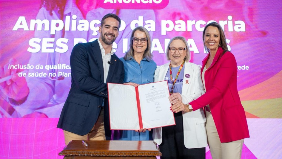 Quatro pessoas posam segurando um documento em frente a um painel rosa com os dizeres “Ampliação da parceria SES RS e IMAMA”. À esquerda está o governador Eduardo Leite, seguido por três mulheres. Todos sorriem para a câmera.