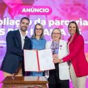 Quatro pessoas posam segurando um documento em frente a um painel rosa com os dizeres “Ampliação da parceria SES RS e IMAMA”. À esquerda está o governador Eduardo Leite, seguido por três mulheres. Todos sorriem para a câmera.