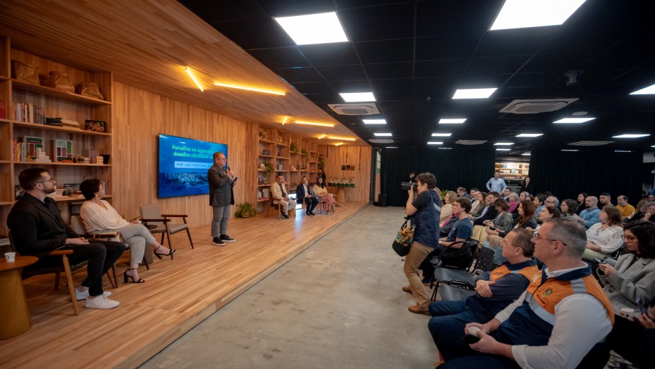 Imagem de um evento profissional realizado em ambiente interno. O local possui paredes revestidas de madeira e estantes com livros e objetos decorativos. No palco, há cinco pessoas sentadas, enquanto uma pessoa está de pé, falando ao microfone. A plateia está disposta em fileiras de cadeiras, voltada para o palco, acompanhando atentamente a apresentação. O espaço tem design moderno, com iluminação de teto e atmosfera organizada, sugerindo uma atividade como seminário, painel de discussão ou workshop.
