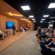 Imagem de um evento profissional realizado em ambiente interno. O local possui paredes revestidas de madeira e estantes com livros e objetos decorativos. No palco, há cinco pessoas sentadas, enquanto uma pessoa está de pé, falando ao microfone. A plateia está disposta em fileiras de cadeiras, voltada para o palco, acompanhando atentamente a apresentação. O espaço tem design moderno, com iluminação de teto e atmosfera organizada, sugerindo uma atividade como seminário, painel de discussão ou workshop.
