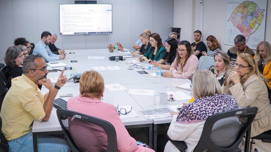 Imagem de uma reunião formal em sala de conferência. Cerca de 16 pessoas estão sentadas ao redor de uma mesa retangular, participando de discussões ou fazendo anotações. O ambiente é bem iluminado, com paredes brancas e luzes no teto. Ao fundo, há uma tela grande exibindo texto, possivelmente parte de uma apresentação ou pauta da reunião. Na parede lateral direita, há um mapa com regiões destacadas em diferentes cores. Os participantes estão vestidos em trajes sociais ou esporte fino e têm documentos e dispositivos eletrônicos sobre a mesa. A imagem retrata um momento de deliberação institucional ou técnica.