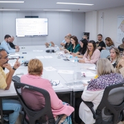 Imagem de uma reunião formal em sala de conferência. Cerca de 16 pessoas estão sentadas ao redor de uma mesa retangular, participando de discussões ou fazendo anotações. O ambiente é bem iluminado, com paredes brancas e luzes no teto. Ao fundo, há uma tela grande exibindo texto, possivelmente parte de uma apresentação ou pauta da reunião. Na parede lateral direita, há um mapa com regiões destacadas em diferentes cores. Os participantes estão vestidos em trajes sociais ou esporte fino e têm documentos e dispositivos eletrônicos sobre a mesa. A imagem retrata um momento de deliberação institucional ou técnica.