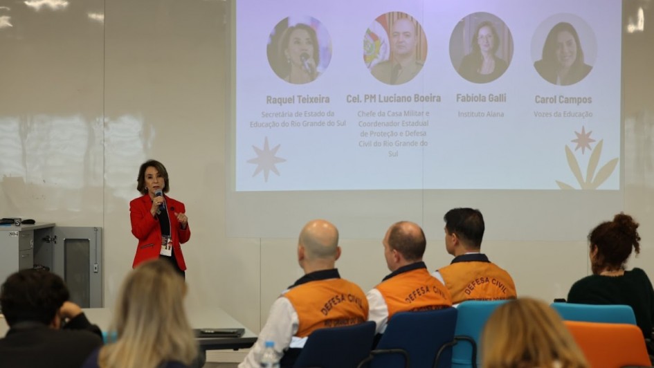 Imagem de uma apresentação institucional em ambiente de conferência. Uma pessoa está em pé, falando ao microfone, vestindo blazer vermelho. Ao fundo, há um slide projetado na parede com o título “ABERTURA INSTITUCIONAL”, que exibe quatro fotos com nomes e cargos:
Raquel Teixeira – Secretária de Estado da Educação do RS
Cel. PM Luciano Boeira – Chefe da Casa Militar e Coordenador Estadual de Proteção e Defesa Civil do RS
Fabiola Galli – Instituto Alana
Carol Campos – Vozes da Educação
Na plateia, algumas pessoas estão sentadas, incluindo participantes com coletes laranja com a inscrição “DEFESA CIVIL” nas costas. O ambiente sugere um evento formal voltado à educação e gestão de riscos climáticos.
