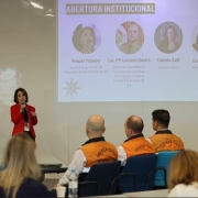 Imagem de uma apresentação institucional em ambiente de conferência. Uma pessoa está em pé, falando ao microfone, vestindo blazer vermelho. Ao fundo, há um slide projetado na parede com o título “ABERTURA INSTITUCIONAL”, que exibe quatro fotos com nomes e cargos:

Raquel Teixeira – Secretária de Estado da Educação do RS
Cel. PM Luciano Boeira – Chefe da Casa Militar e Coordenador Estadual de Proteção e Defesa Civil do RS
Fabiola Galli – Instituto Alana
Carol Campos – Vozes da Educação

Na plateia, algumas pessoas estão sentadas, incluindo participantes com coletes laranja com a inscrição “DEFESA CIVIL” nas costas. O ambiente sugere um evento formal voltado à educação e gestão de riscos climáticos.
