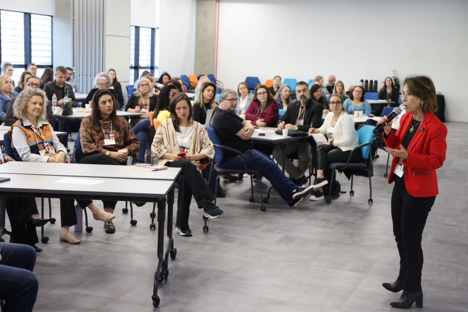Imagem de uma sala de conferência ou formação educacional. Diversas pessoas estão sentadas em mesas, prestando atenção a uma apresentadora em pé, que fala ao microfone e veste blazer vermelho. As mesas estão organizadas de forma que todos os participantes tenham visão clara da apresentação. Ao fundo, há cadeiras coloridas e grandes janelas que permitem a entrada de luz natural, conferindo ao ambiente um aspecto moderno e acolhedor. A cena retrata um momento de capacitação ou evento institucional, com os participantes engajados na atividade.