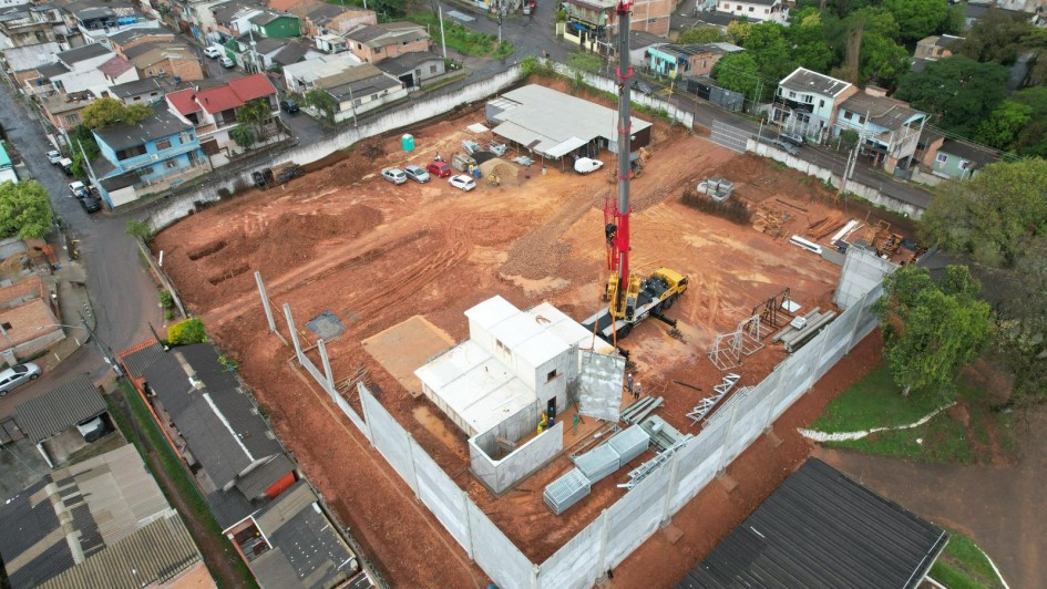 A imagem mostra uma vista aérea de um canteiro de obras em estágio avançado, localizado em uma área urbana cercada por residências. O terreno é amplo, com solo avermelhado, e já conta com muros de concreto. No interior, há estruturas em construção, guindastes e veículos de apoio, além de áreas destinadas a materiais e equipamentos.