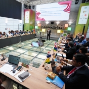 Foto mostra palestrante em pé, em frente a um painel que diz "Semana Fazendária", numa sala com uma grande mesa em formato de U, na qual estão dispostos dezenas de representantes dos estados brasileiros. Na mesa na frente de cada representante há uma pequena bandeira de seus Estados. 