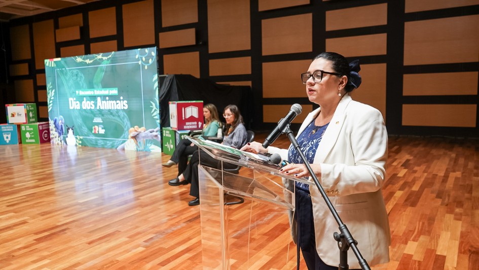 A imagem destaca uma mulher, titular da Sema, que fala ao microfone em um púlpito transparente. Ao fundo, aparecem outras duas mulheres sentadas. Também há um telão com o título do evento: 1º Encontro Estadual pelo Dia dos Animais.