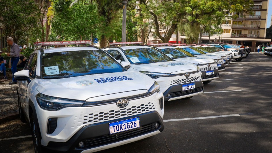 A imagem mostra uma fileira de veículos brancos da marca Toyota estacionados em uma área externa, possivelmente um estacionamento. Os carros estão identificados com a inscrição “Polícia Civil” no capô e possuem sirenes com luzes azul e vermelha no teto. A placa do primeiro veículo da fila é “TOR1G26”. Ao fundo, há árvores, um poste de iluminação e edificações, indicando que o local está em uma área urbana.