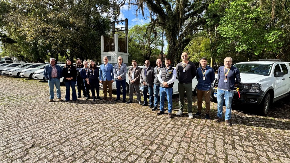 A imagem apresenta um grupo de pessoas posicionado em frente a uma fileira de veículos, em um pátio arborizado. Elas estão alinhadas lado a lado, posando para  a foto de registro de entrega dos automóveis. Uma caminhonete branca está visível à direita, enquanto os demais carros aparecem ao fundo.
