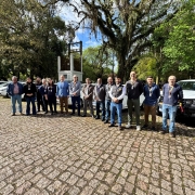 A imagem apresenta um grupo de pessoas posicionado em frente a uma fileira de veículos, em um pátio arborizado. Elas estão alinhadas lado a lado, posando para  a foto de registro de entrega dos automóveis. Uma caminhonete branca está visível à direita, enquanto os demais carros aparecem ao fundo.