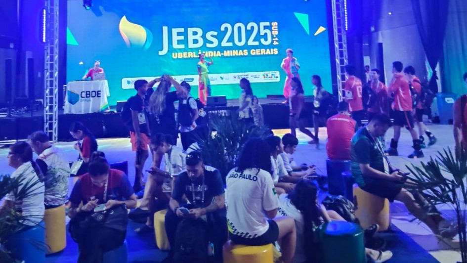 Foto mostra ambiente interno iluminado por luzes coloridas, com jovens sentados em pufes de diferentes cores diante de um palco. No telão ao fundo, aparece o texto “JEBs 2025 Uberlândia - Minas Gerais”. Pessoas circulam e conversam no local.