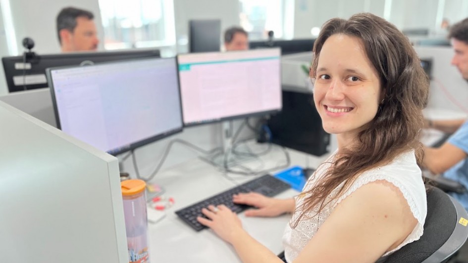 A imagem mostra um ambiente com várias estações de trabalho organizadas em baias. Em primeiro plano, há uma mulher sentada em frente a dois monitores grandes, digitando no teclado. Ao fundo, é possível ver outras pessoas trabalhando em suas respectivas estações, todas equipadas com computadores semelhantes. 