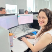 A imagem mostra um ambiente com várias estações de trabalho organizadas em baias. Em primeiro plano, há uma mulher sentada em frente a dois monitores grandes, digitando no teclado. Ao fundo, é possível ver outras pessoas trabalhando em suas respectivas estações, todas equipadas com computadores semelhantes. 