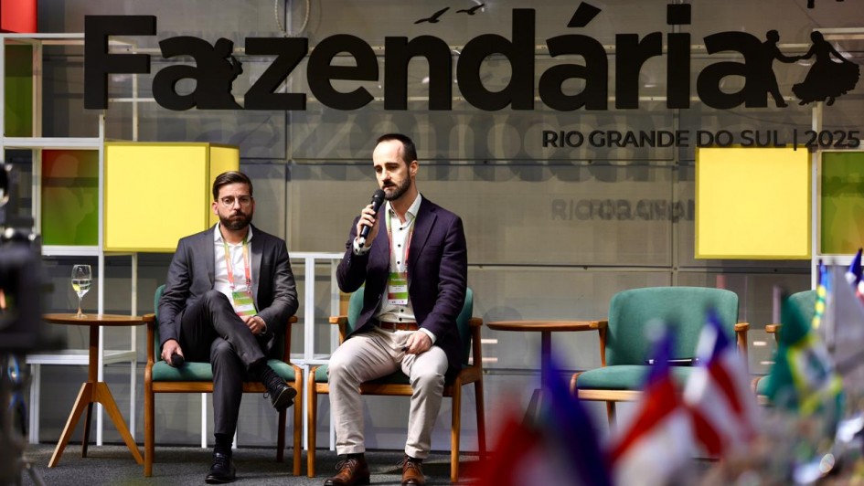 Duas pessoas estão sentadas em cadeiras verdes sobre um palco. Uma delas segura um microfone e parece estar falando, enquanto a outra permanece com as mãos no colo. Ao fundo, há uma grande faixa com os dizeres: “SEMANA Fazendária RIO GRANDE DO SUL 2025”. Na parte inferior da imagem, é possível ver algumas bandeiras desfocadas.