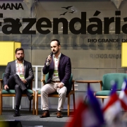 Duas pessoas estão sentadas em cadeiras verdes sobre um palco. Uma delas segura um microfone e parece estar falando, enquanto a outra permanece com as mãos no colo. Ao fundo, há uma grande faixa com os dizeres: “SEMANA Fazendária RIO GRANDE DO SUL 2025”. Na parte inferior da imagem, é possível ver algumas bandeiras desfocadas.