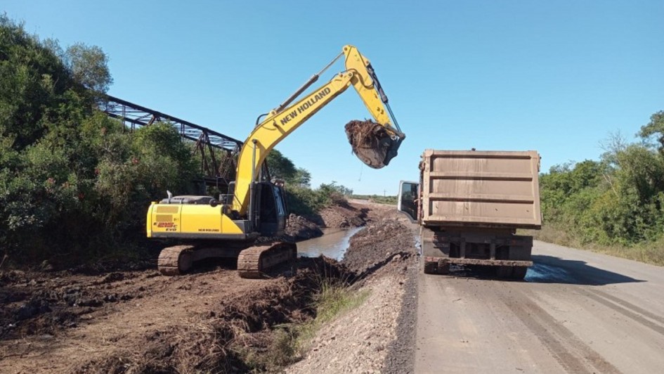 A imagem mostra uma escavadeira hidráulica amarela da marca New Holland operando ao lado de uma estrada asfaltada. A escavadeira está carregando terra e detritos em um caminhão basculante que está estacionado parcialmente sobre a pista. À esquerda, há vegetação densa e uma antiga ponte metálica, parcialmente coberta por árvores. O solo ao lado da estrada está encharcado e com marcas de lama, indicando que está sendo feita uma limpeza ou terraplenagem, possivelmente após enchentes ou deslizamentos. O céu está limpo e azul, indicando um dia ensolarado.