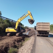A imagem mostra uma escavadeira hidráulica amarela da marca New Holland operando ao lado de uma estrada asfaltada. A escavadeira está carregando terra e detritos em um caminhão basculante que está estacionado parcialmente sobre a pista. À esquerda, há vegetação densa e uma antiga ponte metálica, parcialmente coberta por árvores. O solo ao lado da estrada está encharcado e com marcas de lama, indicando que está sendo feita uma limpeza ou terraplenagem, possivelmente após enchentes ou deslizamentos. O céu está limpo e azul, indicando um dia ensolarado.