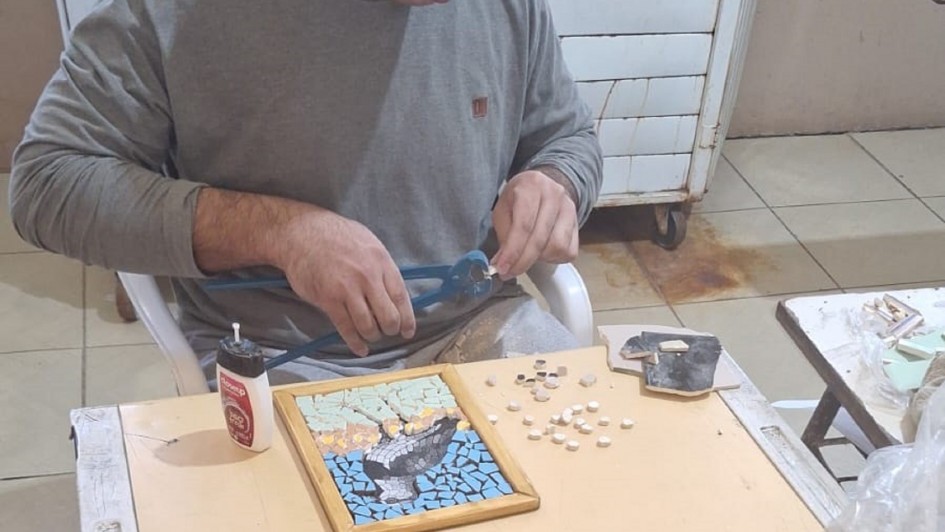 Uma pessoa está sentada à mesa trabalhando em um projeto de arte em mosaico. Ela utiliza uma ferramenta para cortar ou moldar pequenas peças de material, provavelmente azulejos ou pedras. Sobre a mesa, há um mosaico em andamento que representa o desenho de uma ave em voo contra um fundo azul. Diversos pequenos azulejos brancos estão espalhados ao redor da peça. Também é possível ver um frasco de cola e outra ferramenta sobre a mesa. O ambiente é interno, com piso de cerâmica e alguns móveis ao fundo.