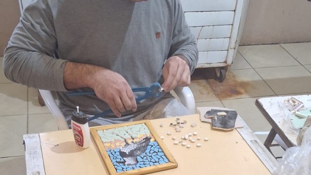 Uma pessoa está sentada à mesa trabalhando em um projeto de arte em mosaico. Ela utiliza uma ferramenta para cortar ou moldar pequenas peças de material, provavelmente azulejos ou pedras. Sobre a mesa, há um mosaico em andamento que representa o desenho de uma ave em voo contra um fundo azul. Diversos pequenos azulejos brancos estão espalhados ao redor da peça. Também é possível ver um frasco de cola e outra ferramenta sobre a mesa. O ambiente é interno, com piso de cerâmica e alguns móveis ao fundo.