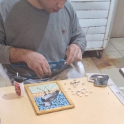 Uma pessoa está sentada à mesa trabalhando em um projeto de arte em mosaico. Ela utiliza uma ferramenta para cortar ou moldar pequenas peças de material, provavelmente azulejos ou pedras. Sobre a mesa, há um mosaico em andamento que representa o desenho de uma ave em voo contra um fundo azul. Diversos pequenos azulejos brancos estão espalhados ao redor da peça. Também é possível ver um frasco de cola e outra ferramenta sobre a mesa. O ambiente é interno, com piso de cerâmica e alguns móveis ao fundo.