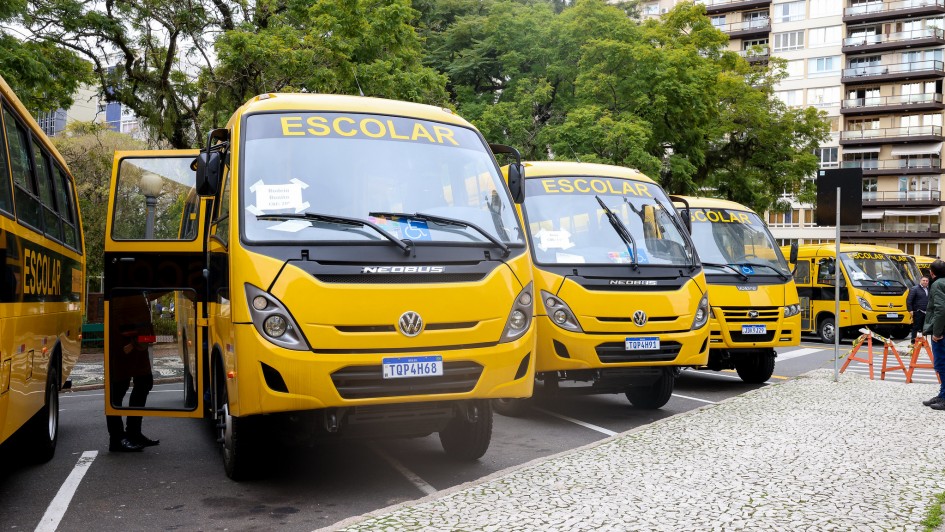 Fila de ônibus escolares amarelos estacionados em via urbana arborizada. Os veículos exibem a palavra “ESCOLAR” na parte superior frontal. Uma pessoa aparece parcialmente ao lado de um dos ônibus, com a porta aberta. Ao fundo, há prédios residenciais.
