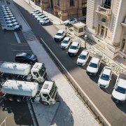 Vista aérea mostra veículos brancos alinhados em frente a um prédio histórico de fachada imponente, com colunas e portas ornamentadas. Entre os veículos, há caminhões-tanque e carros utilitários estacionados paralelamente à calçada.