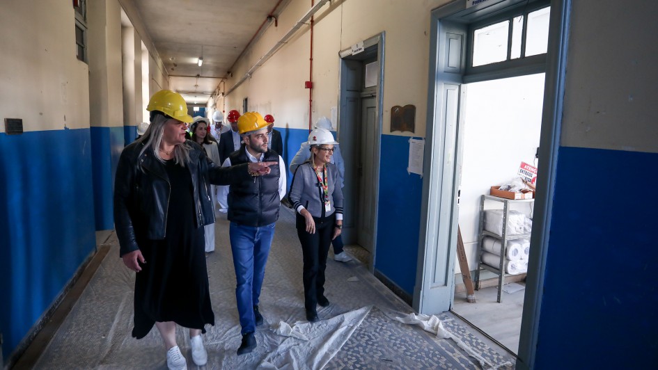 Um grupo de pessoas usando capacetes de segurança caminha por um corredor em obras. As paredes são azuis e bege, e o piso está coberto com lonas de proteção. Uma pessoa à frente aponta para algo, possivelmente explicando ou orientando os demais. Há portas em ambos os lados do corredor, sendo que uma delas, à direita, está aberta e revela uma sala com diversos objetos no interior.