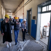 Um grupo de pessoas usando capacetes de segurança caminha por um corredor em obras. As paredes são azuis e bege, e o piso está coberto com lonas de proteção. Uma pessoa à frente aponta para algo, possivelmente explicando ou orientando os demais. Há portas em ambos os lados do corredor, sendo que uma delas, à direita, está aberta e revela uma sala com diversos objetos no interior.