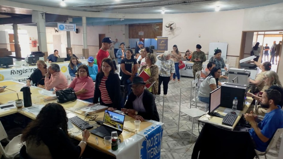 A imagem mostra um ambiente interno, parecido com um salão comunitário, onde está ocorrendo um mutirão de atendimento ao público. Diversas pessoas estão sentadas ou em pé, aguardando ou sendo atendidas em mesas com computadores. O espaço está organizado com mesas distribuídas em fileiras, e há placas com nomes de serviços como "Registro de imóveis", "Projeto Terra" e "Justiça". Algumas pessoas estão com pastas ou documentos nas mãos. Os atendentes estão concentrados nos computadores, realizando cadastros ou tirando dúvidas. O público é composto majoritariamente por adultos e idosos. O ambiente é simples, com piso de cerâmica e teto de madeira, e há ventiladores fixados nas paredes. Ao fundo, há mais pessoas e uma porta aberta por onde entra luz natural. A atmosfera é de organização e prestação de serviços comunitários.