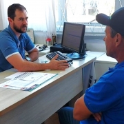Duas pessoas estão sentadas em lados opostos de uma mesa em um ambiente que se assemelha a um escritório. A pessoa à esquerda, vestindo uma camisa azul, gesticula com a mão direita enquanto conversa com a outra. Sobre a mesa, há diversos papéis, um teclado, um mouse e um monitor de computador. A pessoa à direita também usa camisa azul e boné preto. Ao fundo, vê-se uma grande janela com grades e cortinas brancas.