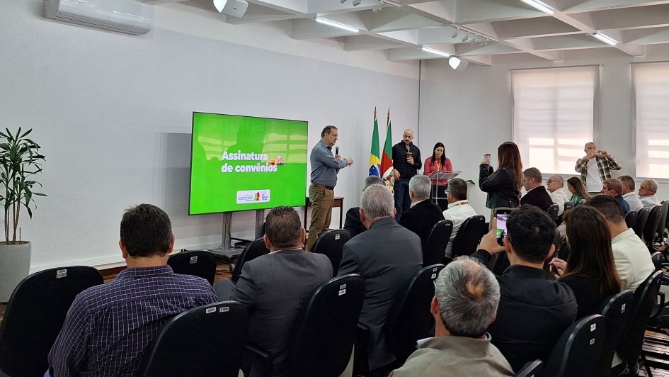 Sala de conferências com várias pessoas sentadas em cadeiras voltadas para um palco. No palco, quatro pessoas estão em pé, sendo que uma delas fala ao microfone. À esquerda, uma tela grande exibe o texto “Assinatura de convênios”. Ao fundo, há três bandeiras posicionadas lado a lado. Algumas pessoas na plateia registram o momento com celulares. O ambiente é bem iluminado e possui janelas grandes no lado direito.