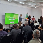 Sala de conferências com várias pessoas sentadas em cadeiras voltadas para um palco. No palco, quatro pessoas estão em pé, sendo que uma delas fala ao microfone. À esquerda, uma tela grande exibe o texto “Assinatura de convênios”. Ao fundo, há três bandeiras posicionadas lado a lado. Algumas pessoas na plateia registram o momento com celulares. O ambiente é bem iluminado e possui janelas grandes no lado direito.