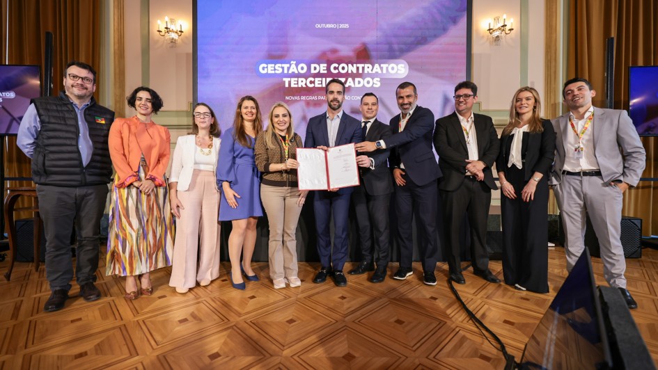 Um grupo de 12 pessoas, formado por homens e mulheres, posa sorridente para uma foto em um ambiente formal, com decoração elegante e piso de madeira trabalhada. Todos estão vestidos com roupas sociais, adequadas a um evento corporativo ou institucional. Ao centro, duas pessoas seguram um documento oficial aberto, emoldurado com capa vermelha, como um certificado ou contrato.
Ao fundo, há uma tela grande com o texto:
"OUTUBRO | 2025
GESTÃO DE CONTRATOS TERCEIRIZADOS
Novas regras para a gestão contratual", indicando que o evento se relaciona com mudanças ou diretrizes em contratos terceirizados. O ambiente parece ser um salão de eventos governamental ou institucional, com cortinas douradas, colunas e lustres de parede.