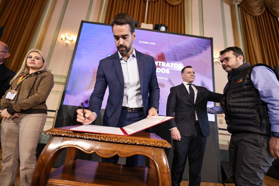 Em um ambiente formal e bem iluminado, um homem de terno azul marinho assina um documento oficial apoiado sobre uma pequena mesa de madeira entalhada, em primeiro plano. Ele segura o documento com uma das mãos e assina com a outra, com expressão concentrada. Ao fundo, uma grande tela digital exibe o título “Gestão de Contratos Terceirizados – Outubro 2025”. À direita, outro homem de terno escuro observa a cena com uma mão no ombro de um terceiro homem que veste colete preto com a bandeira do Rio Grande do Sul. À esquerda, uma mulher loira de expressão séria observa a assinatura. Ela usa calça bege, casaco marrom estampado e crachá.
O local tem cortinas douradas, paredes decoradas e lustres elegantes, reforçando o caráter solene do evento.